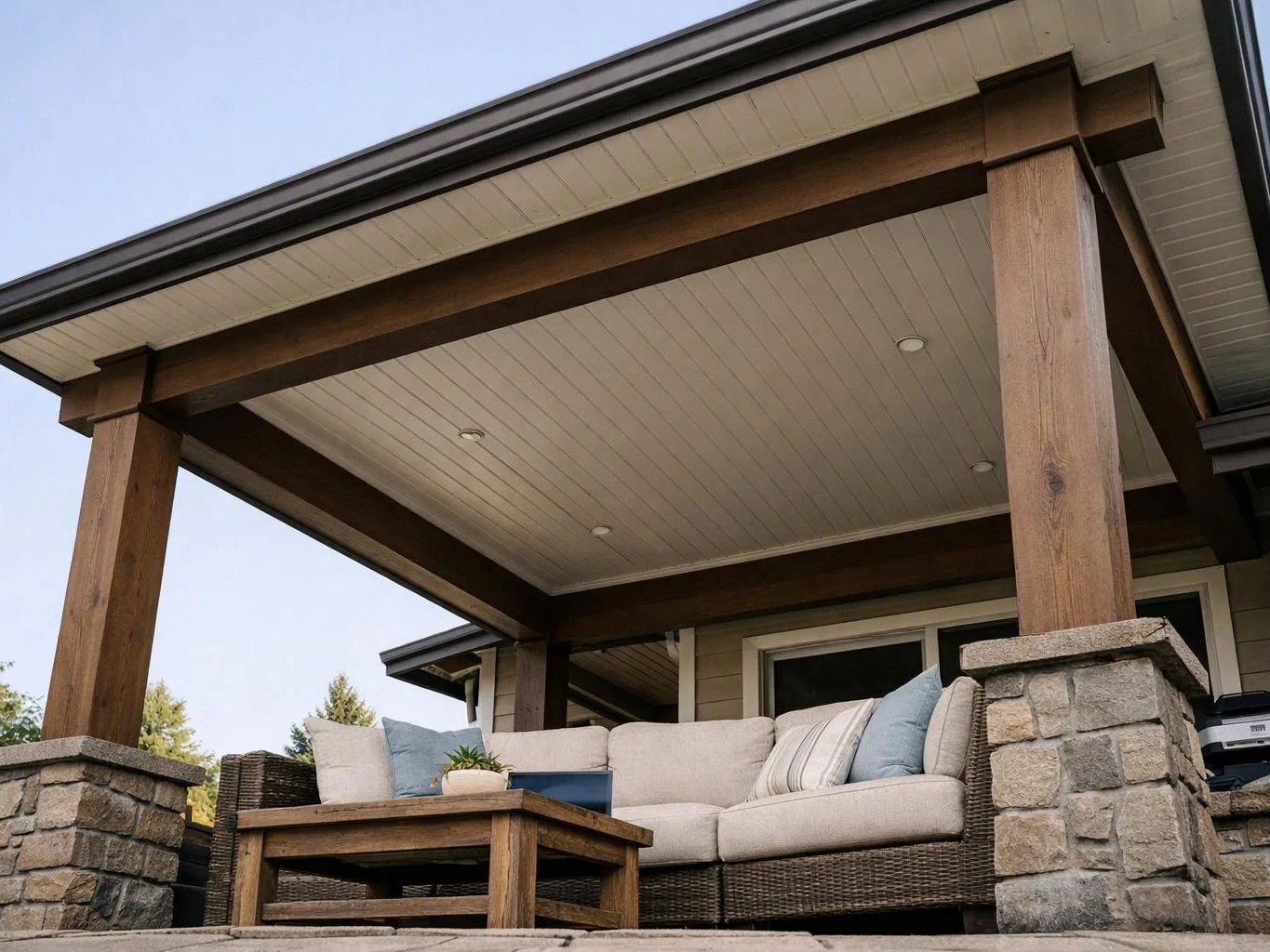 A covered patio shows a wicker sofa, wooden coffee table, and stone columns under a roofed porch