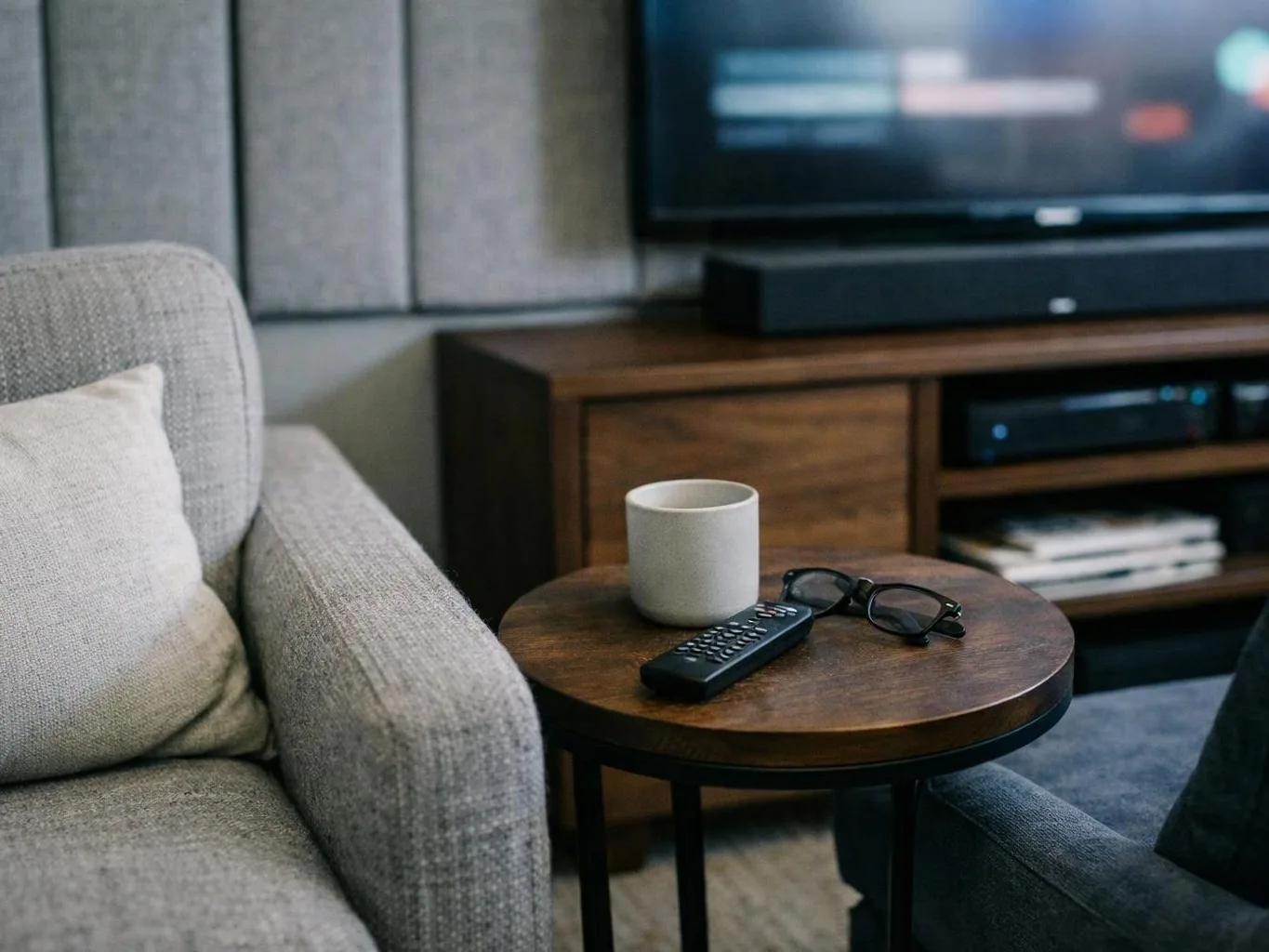 A cozy living room shows a couch, side table with mug, remote, and glasses, and a TV in the background