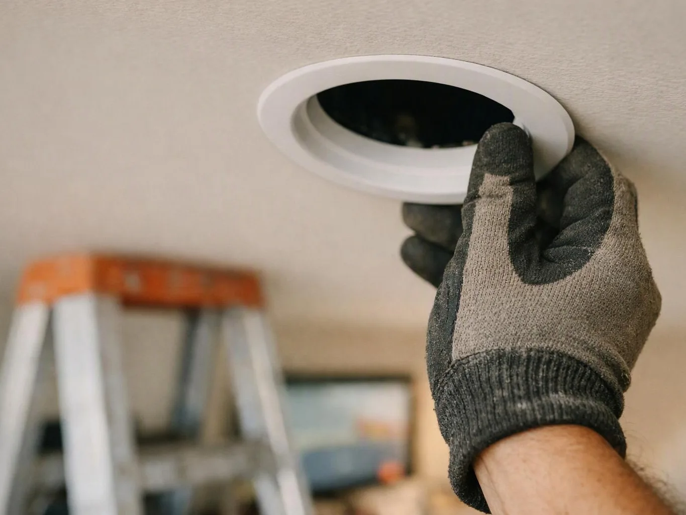 A gloved hand holds a recessed ceiling light trim while a ladder sits blurred in the background