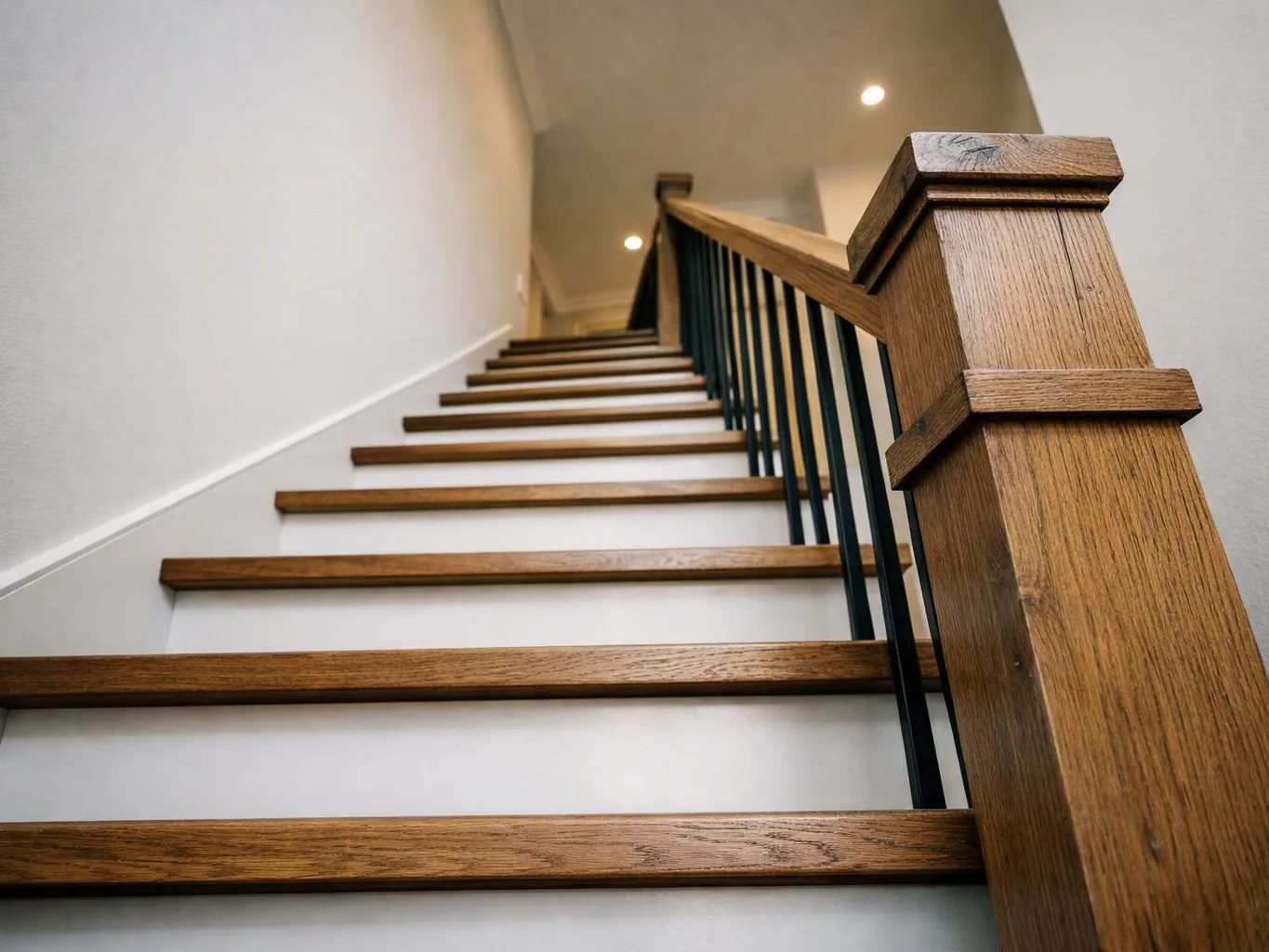 A modern indoor staircase with wooden steps, a dark railing, and white walls rises toward a lit upper landing